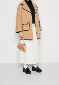 Beżowy płaszcz o fakturze z czarną lamówką, dwie przednie kieszenie, noszony na czarnym golfie. Biała spódnica, czarne masywne mokasyny oraz mała beżowa torebka.