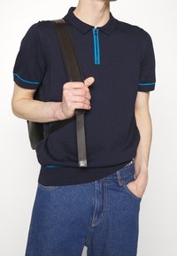 Námořnická polokošile s krátkým rukávem má límec na zip s tyrkysovými akcenty. V kombinaci se světle modrými džínami a černou taškou na rameno.
