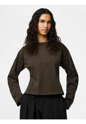 Ung person med langt krøllete hår, iført en mørkebrun langermet skjorte og svarte plisserede bukser, stående med hendene i lommene.