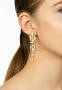 Orecchini d'oro pendenti con design a forma di foglia, caratterizzati da gemme trasparenti e una finitura testurizzata, che si abbinano a un'acconciatura elegante.