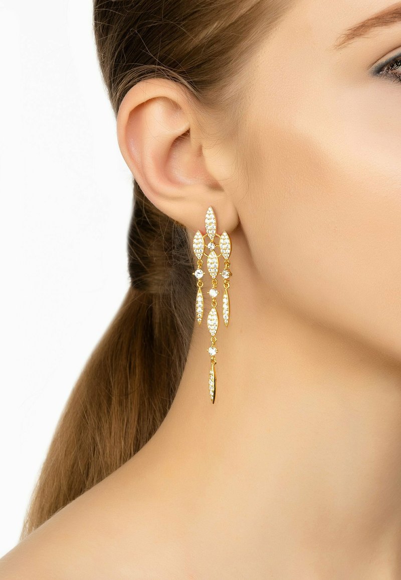 Orecchini d'oro pendenti con design a forma di foglia, caratterizzati da gemme trasparenti e una finitura testurizzata, che si abbinano a un'acconciatura elegante.