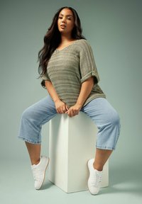 Suéter de punto suelto en color verde oliva con escote en V, combinado con jeans recortados de color azul claro y zapatillas blancas, sobre una modelo sentada en un pedestal blanco.