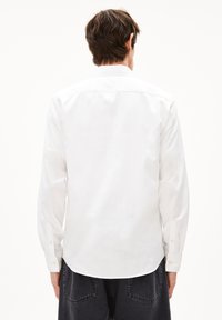 Mann mit kurzen braunen Haaren, der ein langärmeliges weißes Hemd und schwarze Jeans trägt, steht mit dem Rücken zu einem weißen Hintergrund.
