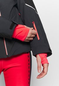 Zwarte jas met feloranje accenten en een ritssluiting op de mouw. Rode nauwsluitende broek met een zwart zijpaneel en stikseldetails.