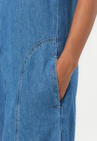Tuta in denim blu con tasca laterale e dettagli di cucitura a vista. Il tessuto appare liscio con una finitura leggermente texturizzata.