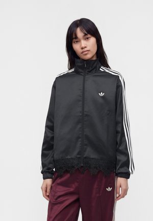 Γυναίκα που φοράει μαύρο αθλητικό μπουφάν Adidas με λευκές ρίγες και δαντελωτή επένδυση, συνδυασμένο με μαρονού χρώματος αθλητικό παντελόνι Adidas.