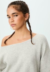 Hellgrauer, schulterfreier Pullover aus weichem Strickstoff mit auffälliger Textur. Das Modell hat zwei geflochtene Zöpfe und große goldene Creolen.
