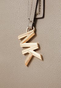 Sac à main en cuir texturé beige avec un pendentif logo métallique doré présentant des formes angulaires, attaché par une courte bandoulière.