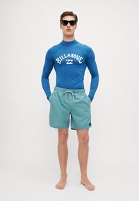 Zils garām piedurknēm pārklājums ar "BILLABONG" logo, apvienots ar gaiši varavīksnenes peldbiksēm un bežām aukliņām. Valkā melnas saulesbrilles.