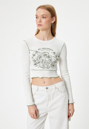 Valkoinen ribattu pitkähihainen croptop, jossa on vihreä ommel, kukkakuva ja teksti: "Ole Luova." Pehmeä pinta ja aaltoileva helma.