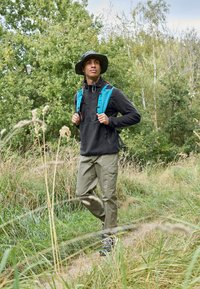 Outdoor udstyr med en sort fleece trøje, olivengrøn cargo-bukser, en blå rygsæk og en bredskygget hat, sat mod en grøn naturbaggrund.
