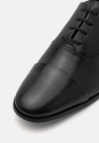 Zapato de vestir de cuero negro con un diseño elegante, textura suave, puntera redondeada y cierre de cordones tradicional con un sutil detalle de costura.