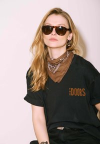 Femme blonde portant des lunettes de soleil écaille de tortue, un bandana brun à motifs, un t-shirt noir des Doors et des boucles d'oreilles pendantes sur fond clair.