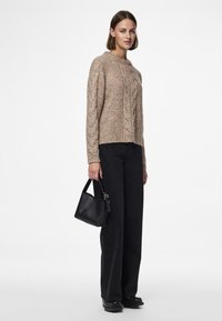 Beiger Strickpullover mit Zopfmuster, lockere Passform, Rundhalsausschnitt, kombiniert mit hoch taillierten schwarzen weit geschnittenen Jeans und einer kleinen schwarzen Tasche.