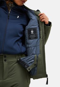 Olivengrüne Jacke mit einem marineblauen Innenfutter, ausgestattet mit einer gesteppten Textur, einem Logo-Patch und einer Reißverschlusstasche. Die Details umfassen kontrastierende Einsätze.