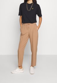 Zwarte t-shirt met een geel logo, beige broek met een elastische tailleband en witte sneakers. De broek heeft zijzakken en een losse pasvorm.