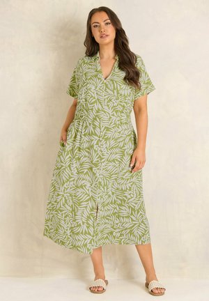 Vrouw staand in een groene midi-jurk met witte bladpatronen, korte mouwen, een kraag en instapsandalen met bloemmotief.