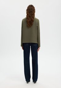 Olivgrüne, kurz geschnittene Jacke mit langen Ärmeln, aus strukturiertem Stoff, kombiniert mit dunkelblauen, weit geschnittenen Jeans. Schlichtes Design, keine sichtbaren Muster oder Akzente.