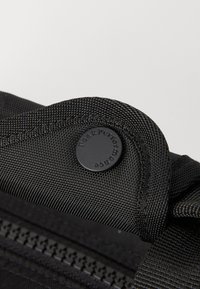 Black fabric bag featuring a textured design, a round black button with "Peak Performance" embossed, and a visible zipper detail.
