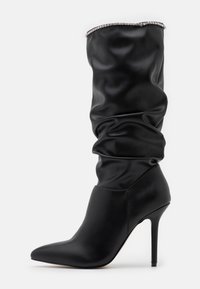 Bota negra hasta la rodilla hecha de cuero liso, con un diseño holgado, punta afilada y tacón alto fino. Destaca un adorno de cristales en la parte superior.