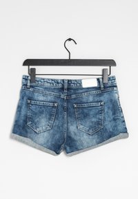 Shorts en denim bleu avec des ourlets retroussés pendus à un cintre noir, montrant des poches arrières et une étiquette blanche sur la ceinture.