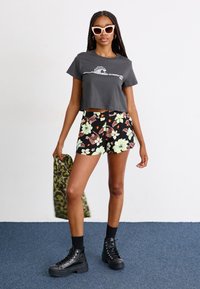 Žena v černém crop topu s vlnovým potiskem, květinové šortky, černé boty, drží zelenou bundu s leopardím vzorem a bílé sluneční brýle ve tvaru kočičích očí.