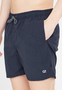 Mörkblå badshorts i lätt material, med elastisk midja och sn örning, sidofickor och en liten vit logotyp.