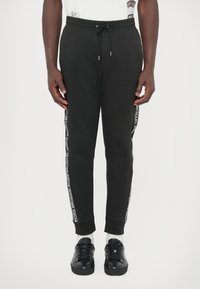 Czarne spodnie dresowe z mieszanki bawełny, z elastycznym pasem ze sznurkiem, zwężanym krojem i taśmą z logo na bocznych szwach. Połączone z czarnymi sportowymi butami.