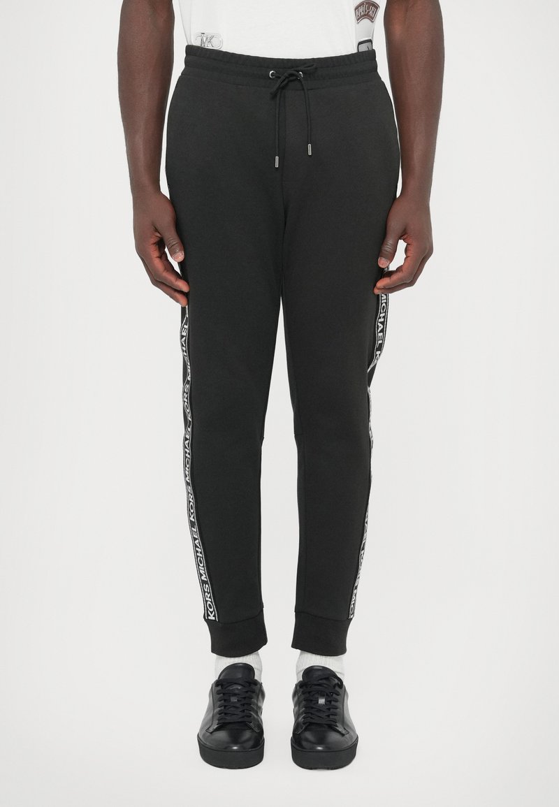 Czarne spodnie dresowe z mieszanki bawełny, z elastycznym pasem ze sznurkiem, zwężanym krojem i taśmą z logo na bocznych szwach. Połączone z czarnymi sportowymi butami.