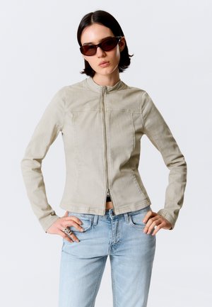 Femme portant une veste beige zippée, un jean bleu clair, des lunettes de soleil foncées et des bagues argentées à la main gauche, sur un fond uni.
