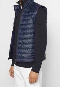 Marinblå ärmlös vadderad väst med quiltad design, dragkedjeöppning och sidofickor, tillverkad av glansigt, lätt material.