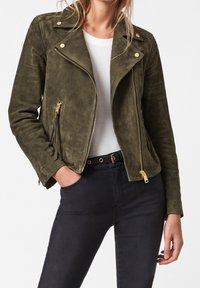 Zielono-oliwkowa skórzana kurtka motocyklowa z złotymi elementami, skośnym zapięciem na zamek i zapinanymi kieszeniami bocznymi. Noszona na białym topie.