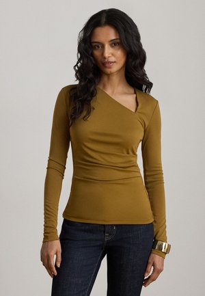 Femme aux longs cheveux foncés portant un haut asymétrique vert olive à manches longues et un jean foncé, posant devant un fond uni.