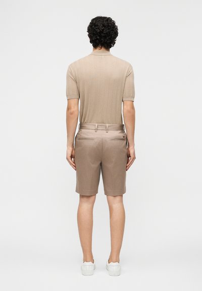 Άνδρας με σγουρά σκούρα μαλλιά, που φοράει μπεζ πόλο με κοντά μανίκια και υφή, ταιριαστό μπεζ σορτς και λευκά αθλητικά παπούτσια, στέκεται με την πλάτη γυρισμένη σε απλό φόντο.