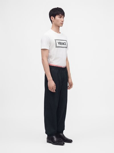 Νέος άνδρας στέκεται φορώντας λευκό t-shirt Versace, μαύρο άνετο παντελόνι με λεπτομέρεια κόκκινης ζώνης και μαύρα δερμάτινα παπούτσια σε απλό φόντο.