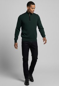 Suéter de punto acanalado verde con cuello de cremallera, combinado con jeans oscuros y botines negros. El suéter tiene detalles en negro en los hombros.