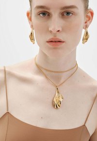 Joyería de oro con formas fluidas y onduladas. Incluye aretes a juego y un collar con colgante, todo sobre un fondo neutro.