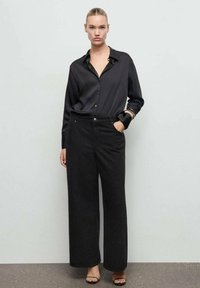 Camisa negra de satén con botones y cuello combinada con pantalones anchos negros de textura y sandalias de tacón negras. Pulsera dorada como acento.