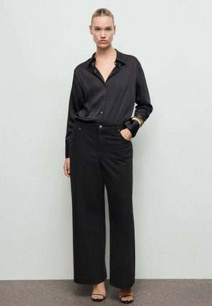 Camisa negra de satén con botones y cuello combinada con pantalones anchos negros de textura y sandalias de tacón negras. Pulsera dorada como acento.
