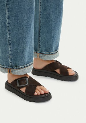 Feet wearing dark brown suede sandals with thick black soles and buckle straps, paired with rolled-up blue denim jeans.