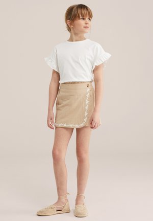 Mlada djevojka stoji, nosi bijelu bluzu s naboranim rukavima, bež vezanu suknju s vezom i bež tkanine cipelama vezanim oko gležnja.