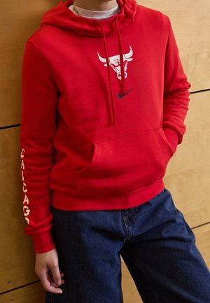 Osoba w czerwonej bluzie z kapturem Chicago Bulls z białym logo i napisem na rękawie, w niebieskich dżinsach, stojąca na tle drewnianej ściany.