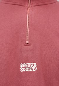 Gros plan sur un sweat-shirt à col quart zippé rose poudré avec un logo "Kassiko Society" brodé en blanc sur la poitrine.