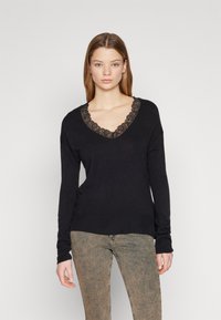 Zwarte trui met een V-hals en een gedetailleerde kantafwerking. Gebreide textuur, lange mouwen en een relaxed fit. Gecombineerd met lichtbruine jeans.