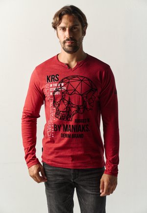 Hombre con camisa roja de manga larga con calavera geométrica y texto en negro, combinada con vaqueros gris oscuro, de pie frente a un fondo claro y liso.