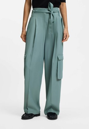 Kvinde iført højtaljede, vide salviegrønne cargobukser med bundet bælte og klaplommer, kombineret med sorte sko og en sort top.