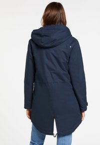 Mujer vistiendo un abrigo parka con capucha de color azul marino, con una abertura en la parte posterior, combinado con jeans de un azul claro, de espaldas a la cámara sobre un fondo blanco.