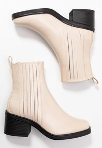 Bottines en cuir synthétique beige avec un talon carré, présentant des détails de couture verticale et deux languettes de tirage pour un enfilage facile.