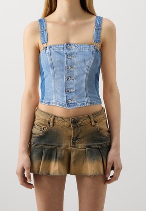 Frau, die ein ärmelloses hellblaues Jeans-Cropped-Top mit Knöpfen und einen braun-schwarz verblassten plissierten Minirock trägt, stehende mit entspannten Armen.