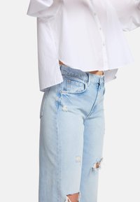 Hvid skjorte med knaplukning, kort længde og brede ærmer, parret med lyseblå slidte jeans med huller og en afslappet pasform.